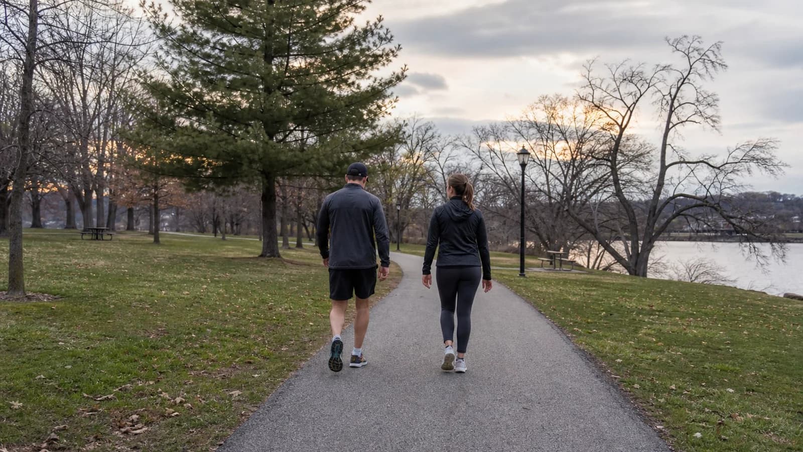 Two people walking on a park path near water at dusk